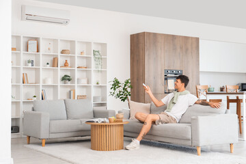 Young man on sofa turning on air conditioner at home