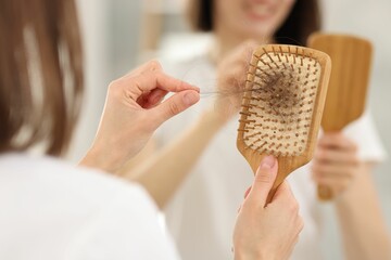 Woman taking her lost hair from brush near mirror indoors, closeup. Alopecia problem