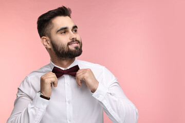 Portrait of smiling man adjusting bow tie on pink background. Space for text