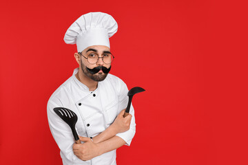 Professional chef with funny artificial moustache holding kitchen utensils on red background. Space...