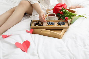 Tasty breakfast served in bed. Woman with tea, desserts, gift box and flowers at home, closeup