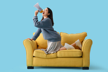 Young woman shouting into megaphone on yellow sofa against blue background