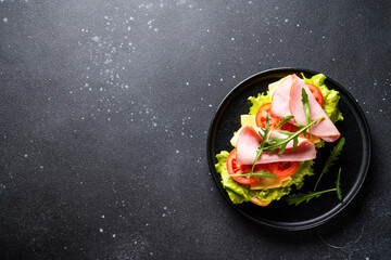 Ciabatta sandwich with lettuce, cheese, tomatoes and ham on black background. Flat lay with copy space.