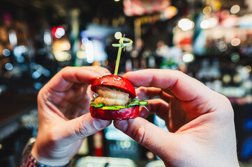 Close-up of hands holding a creative gourmet mini burger with fresh ingredients and a toothpick,...