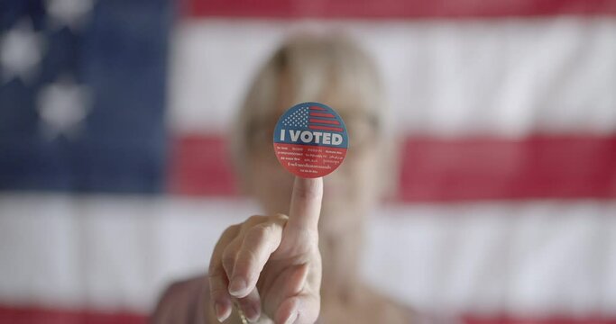 Elderly woman voter holds up "I voted" sticker on finger, in front of her face. Rack focus from face to sticker. United States flag on wall in background. 4K DCI