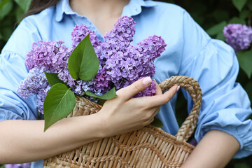 Beautiful young woman holding wicker bag with bouquet of lilac flowers in park on spring day,...