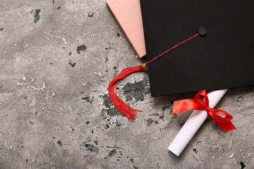 Diploma with red ribbon, book and graduation hat on grey grunge background