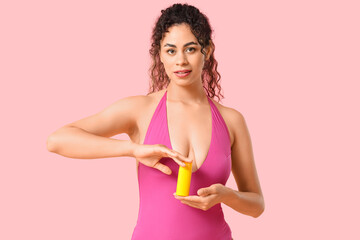 Beautiful young African-American woman in swimsuit with bottle of sunscreen cream on pink background