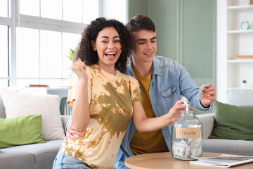 Happy young couple putting dollar banknotes into glass jar at home