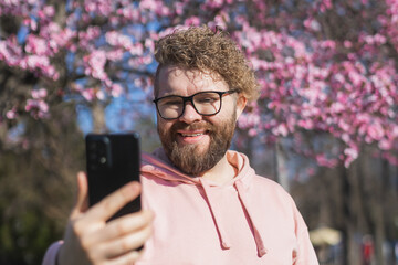 Fototapeta premium Happy curly man takes selfie against backdrop of flowering tree in spring for his internet communications. Weekend and social networks concept