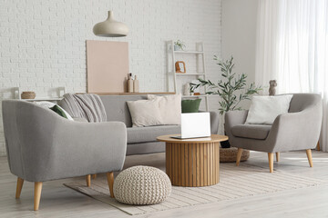 Stylish interior of living room with cozy sofa, armchairs, coffee table, laptop and olive tree