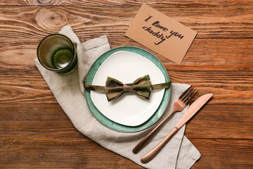Festive decorated table setting with greeting card and bowtie on brown wooden background. Father day celebration concept