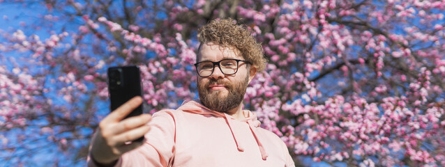 Spring day banner. Bearded man in pink shirt talking by phone. Spring pink sakura blossom. Handsome...