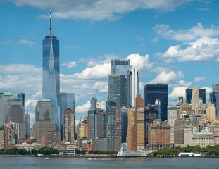 New York, NY, USA - August 1, 2023: One World Trade Center above other skyscrapers in financial district under blue cloudscape. Green battery park and its authority office building with short green ti