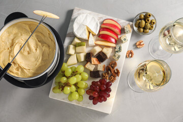 Fork with piece of apple, melted cheese in fondue pot, wine and snacks on grey table, flat lay