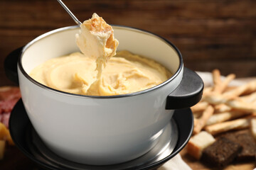 Dipping piece of bread into fondue pot with melted cheese on blurred background, closeup