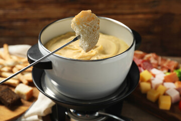 Fork with piece of bread, melted cheese in fondue pot and snacks on table, closeup