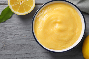 Delicious lemon curd in bowl, fresh citrus fruit and green leaf on grey wooden table, top view....