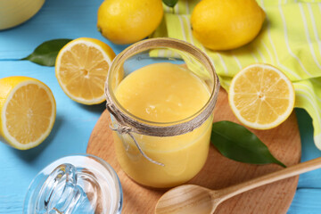 Delicious lemon curd in glass jar, fresh citrus fruits, green leaves and spoon on light blue wooden table