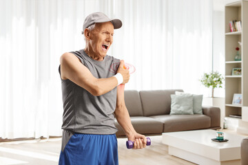 Elderly man in sportswear exercising at home in a living room and holding his shoulder