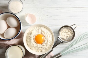 Flour with yolk in bowl and other ingredients for dough on white wooden table, flat lay