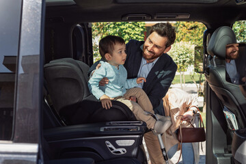 A stylish dad puts his son in a car seat and fastens his seat belt in a minibus