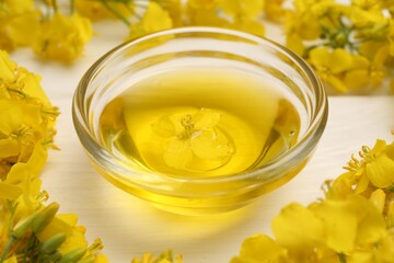 Rapeseed oil in glass bowl and beautiful yellow flowers on white wooden table, closeup