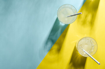 Two glasses of lemonade with a straw and the shadow of another glass on light blue and yellow background with copy space. 