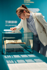 Thoughtful young Caucasian woman wearing glasses and looking at exhibition. Concept of Museum Day.