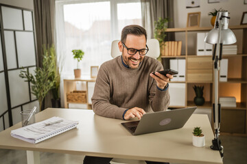 Adult caucasian man at office sit hold mobile phone and work on laptop