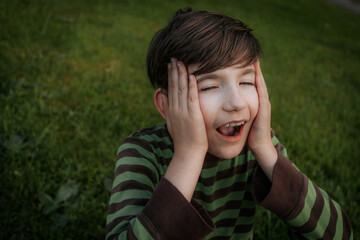 A young boy in a striped shirt sits on the grass, holding his face in surprise with an open mouth. The green background adds a natural, vibrant setting to the playful expression.