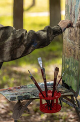 An artist is immersed in capturing the essence of nature, painting en plein air with an array of brushes and oil paints. The scene is set with an easel, palette, and red paintbrush holder