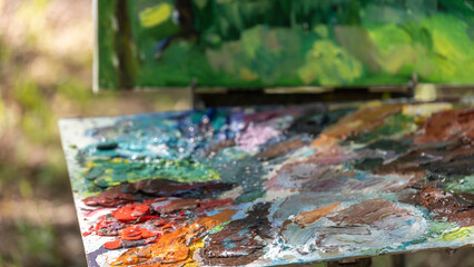 An artist's wooden palette with vibrant oil paint colors sits in focus against a blurred natural landscape, capturing the essence of painting en plein air.