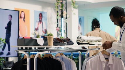 African american employee working in SH clothing shop, placing simple stylish shirts on racks....