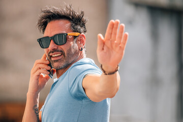 attractive young man in sunglasses talking on mobile phone and waving on the street in summer outdoors