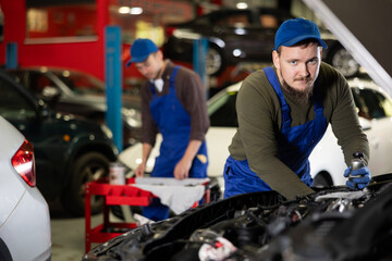Young guy mechanic in uniform repairs under hood of car in car service station