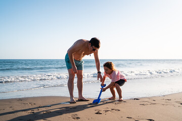 Adult male and young child engage in playful activities with sand toys on a sunny beach, creating...