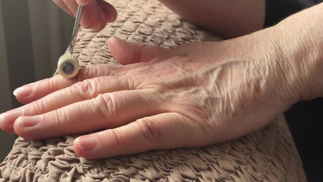 An unknown elderly woman sitting in an armchair makes herself a hand massage with her hand with a massager in the form of a needle-shaped silver roller. Health maintenance and Promotion Program