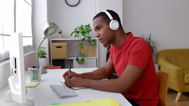 Young African American man sitting at desk working on laptop taking notes in notebook. Gen z boy studying online, watching webinar, listening webinar course. Education and e-learning concept.