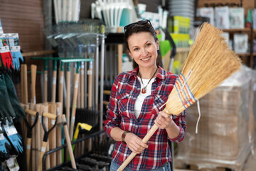 Adult woman shopper in casual clothes choosing broom in household store..