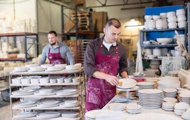 Attentive experienced male ceramicist in maroon apron working in artisanal workshop, using sponge to clean pottery plates before glazing process..