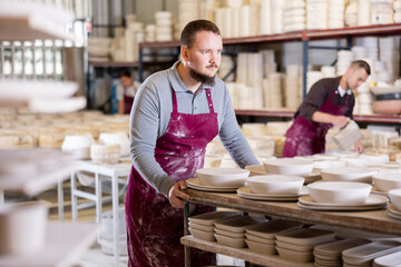 Young male worker in uniform moves rack with ceramic dishes blanks at factory