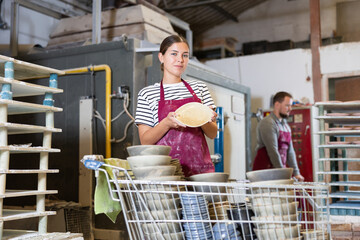 Enthusiastic young female ceramicist in maroon apron holding up freshly handcrafted plates,...