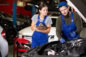 Woman with documents looks at guy mechanic repairs under hood of car in car service station