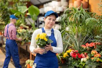 Young female saleswoman in uniform holding pot of begonia in flower shop..