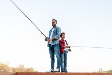 African American man and child sitting on a wooden pier holding fishing rods on the river, dad...