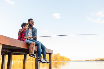 African American man and child sitting on a wooden pier holding fishing rods on the river, dad...