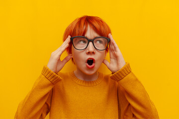 surprised red-haired child boy with glasses looks away on yellow isolated background, shocked...