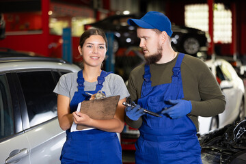Young guy and young woman mechanics in uniform posing in car service station