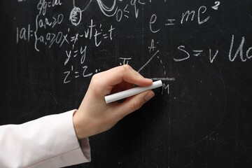 Teacher writing physical formulas with chalk on black chalkboard, closeup